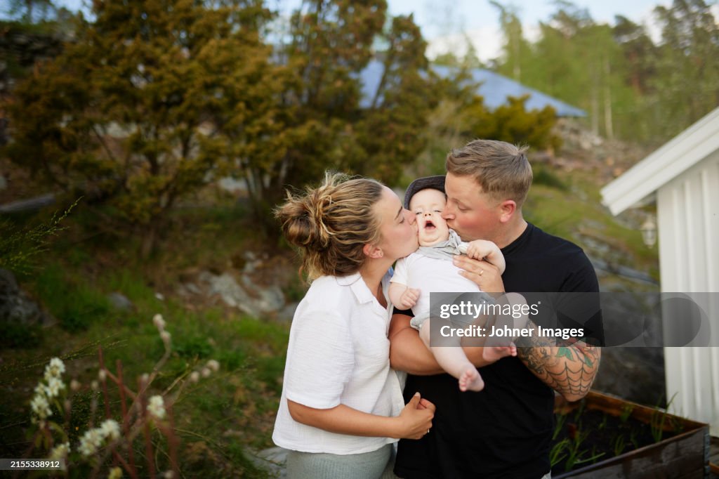 Young parents kissing toddler son while standing together at back yard