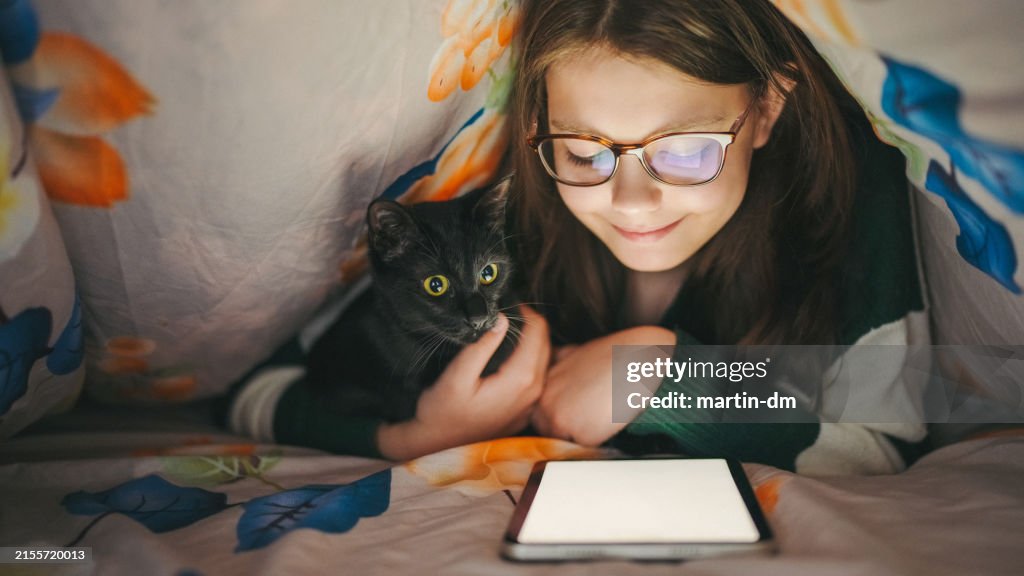 Girl with cat studying on tablet under a blanket