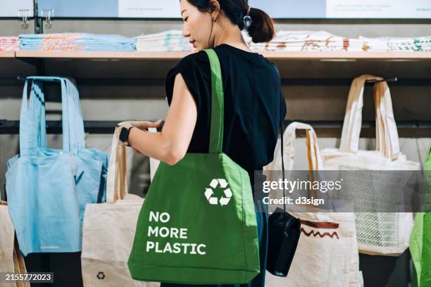 young asian woman carrying a green cotton reusable shopping bag with text no more plastic with a recycling symbol shopping in a store. environmental protection. responsible and ethical shopping. zero waste. sustainable lifestyle. eco-friendly bags concept - bolsa reutilizable fotografías e imágenes de stock