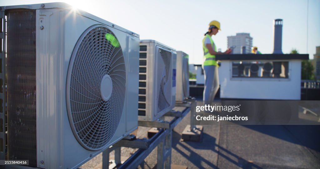 Wartungsarbeiter oder Ingenieur mit Schutzhelm und Warnweste, die den Lüfter der Klimaanlage im Freien untersuchen und Notizen machen