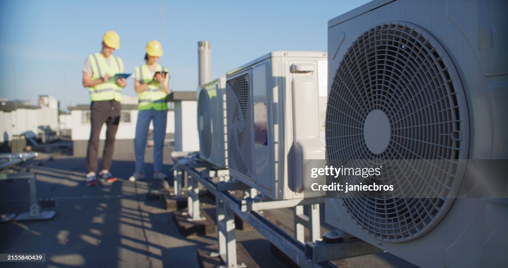 Wartungsarbeiter oder Ingenieure mit Schutzhelmen und gelben Warnwesten, die gemeinsam den Lüfter der Klimaanlage im Freien untersuchen und Notizen machen