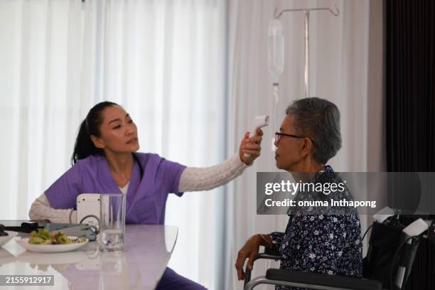 mulher idosa em uma casa de repouso sentada enquanto assistente de saúde toma a temperatura com um termômetro infravermelho na testa. atenção especializada ao idoso aposentado. - ultrasonografia médica equipamento de monitoramento - fotografias e filmes do acervo
