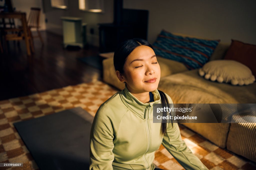 Young Asian woman meditating in living room. Indoor lifestyle portrait. Self-care and mindfulness concept.