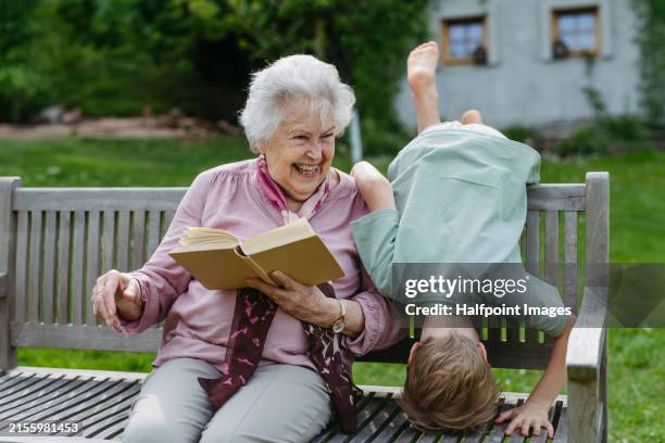 portrait of young boy spending time outdoor with grandmother, doing silly things while she sitting on bench in the garden. grandson enjoying together time with elderly grandma. - comedian stock pictures, royalty-free photos & images