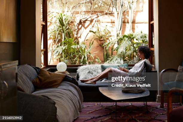 woman using smart phone sitting by window and sofa - boho-stil stock-fotos und bilder