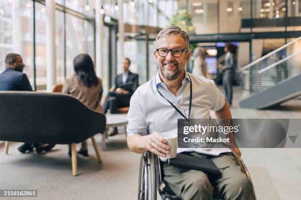 portrait of happy businessman with disability holding coffee cup while sitting on wheelchair at convention center - rolstoel stockfoto's en -beelden