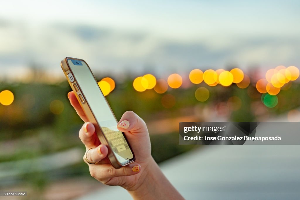 La main d’une jeune femme tenant et utilisant son iPhone sur un fond urbain crépusculaire.