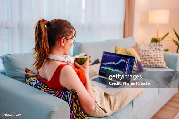 joven empresaria se sienta en el sofá con los pies en alto, taza de café en mano, revisando el gráfico de precios de bitcoin o intercambio en un intercambio digital en su teléfono celular mientras investiga en el monitor de una computadora portátil - opciones fotografías e imágenes de stock