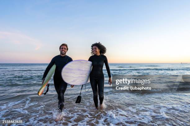 vielfältiges paar mit surfbrettern am strand sonnenuntergang - neoprenanzug stock-fotos und bilder