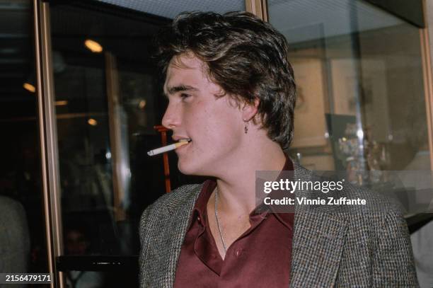 American actor Matt Dillon, who wears a grey tweed blazer over a maroon shirt, and a crucifix earring, a cigarette in his mouth as he attends an...
