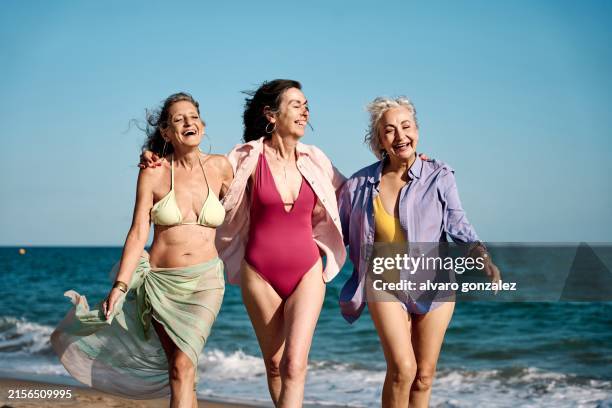 three mature women walking and laughing together on the beach - badkläder bildbanksfoton och bilder