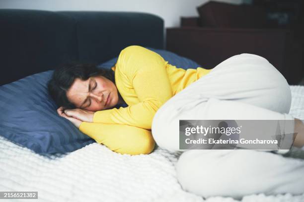 woman lying on bed with hand on forehead. wearing yellow cardigan and blue shirt. relaxed and thoughtful. - brechreiz stock-fotos und bilder