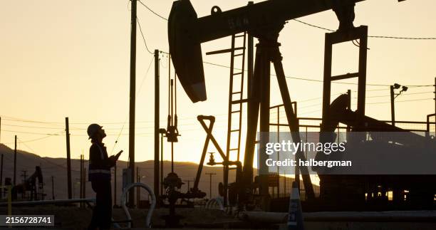 oilfield worker looking up to pump jack with sunset in background - oil pump stock pictures, royalty-free photos & images