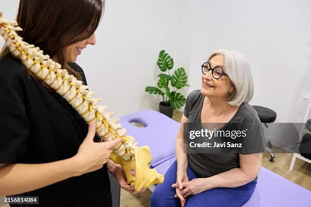 a young female physiotherapist uses a model of the human spine to explain spinal anatomy to an older woman in a physiotherapy clinic - osteoporose imagens e fotografias de stock