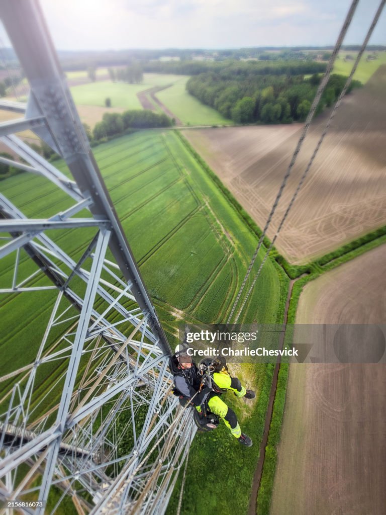 Industrial climber abseiling down from blade of wind turbine on the ropes. Agricultural fields, paths under and sun behind. Lattice tower. Shot from up.