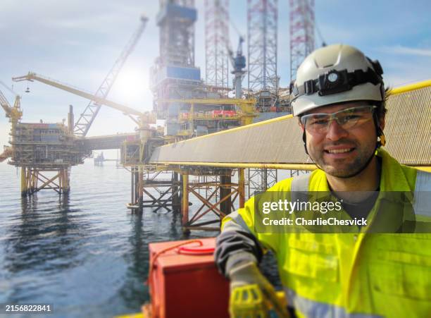 the professional manual worker is standing in front of an oil platform (oil rig) in the north sea at sunrise, smiling. behind him, there's a bridge connecting the platform and the dramatic sky, with the sunrise in the background. he is wearing ppe. - oljeplattform bildbanksfoton och bilder
