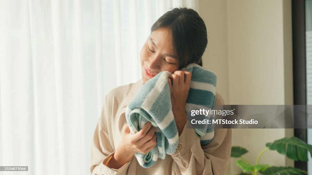 Beautiful young Asian woman smelling clean clothes and smiling while doing laundry at home. Activity of daily living at house.