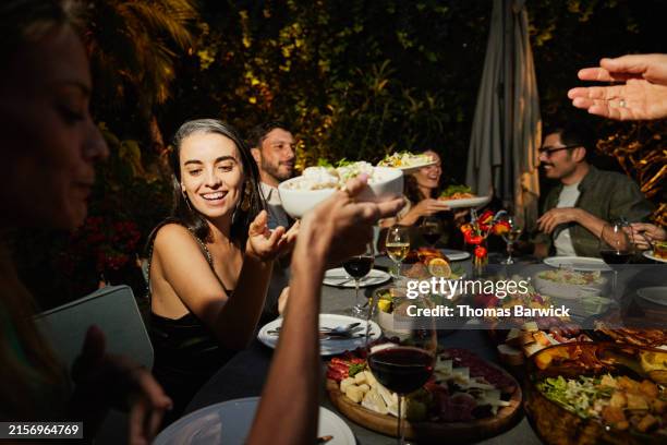 medium shot friends passing plates of food during rooftop dinner party - cena con amigos fotografías e imágenes de stock