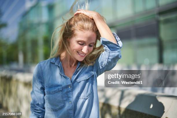 smiling woman with hand in hair and standing near retaining wall - denim shirt stock pictures, royalty-free photos & images