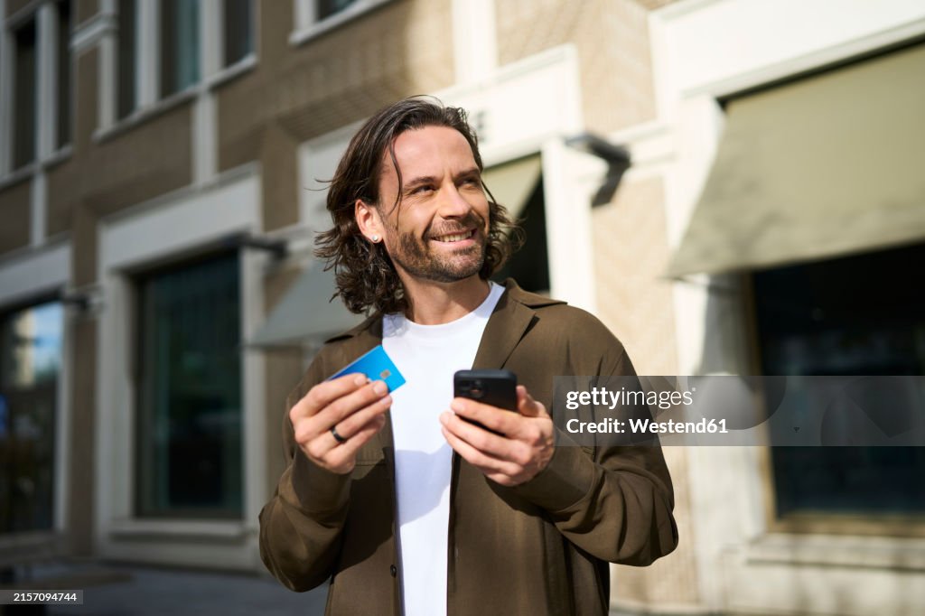 Smiling handsome man using smart phone doing online shopping through credit card on sunny day