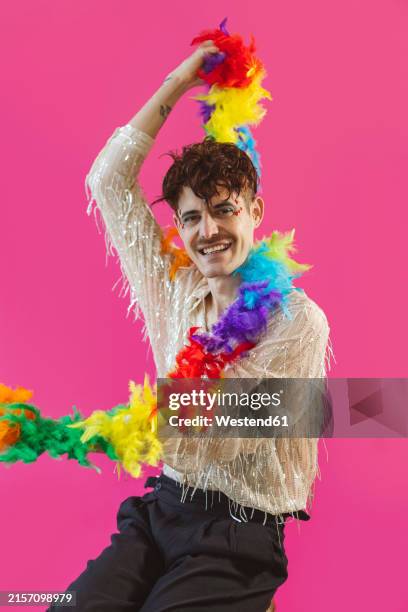 happy non-binary person sitting with rainbow feather boa against pink background - boa persoonlijk accessoire stockfoto's en -beelden