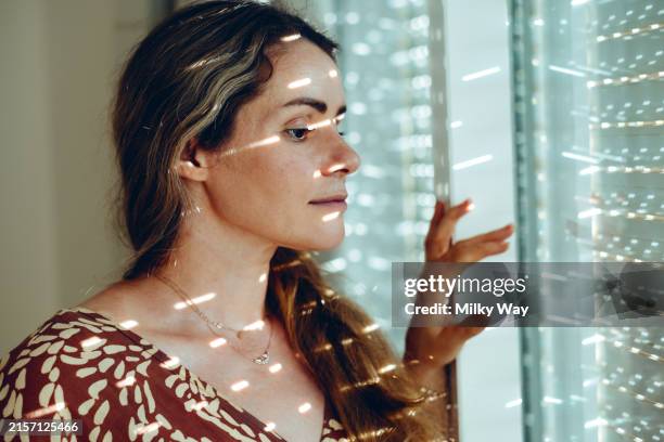 woman gazing thoughtfully through window blinds, with natural light casting striped shadows on her face. - buren stockfoto's en -beelden