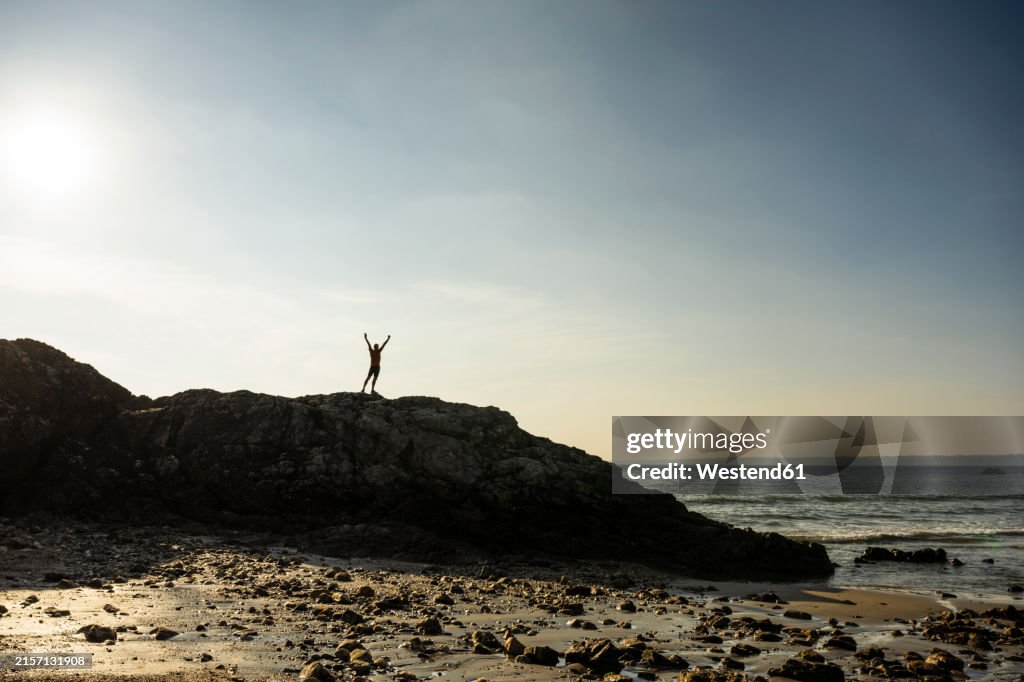 Man with arms raised standing on rock near sea at sunrise