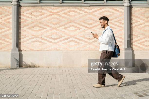 side view of middle aged latin man leaving work dressed in casual clothes sending a text message and smiling - crossbody bag stock pictures, royalty-free photos & images