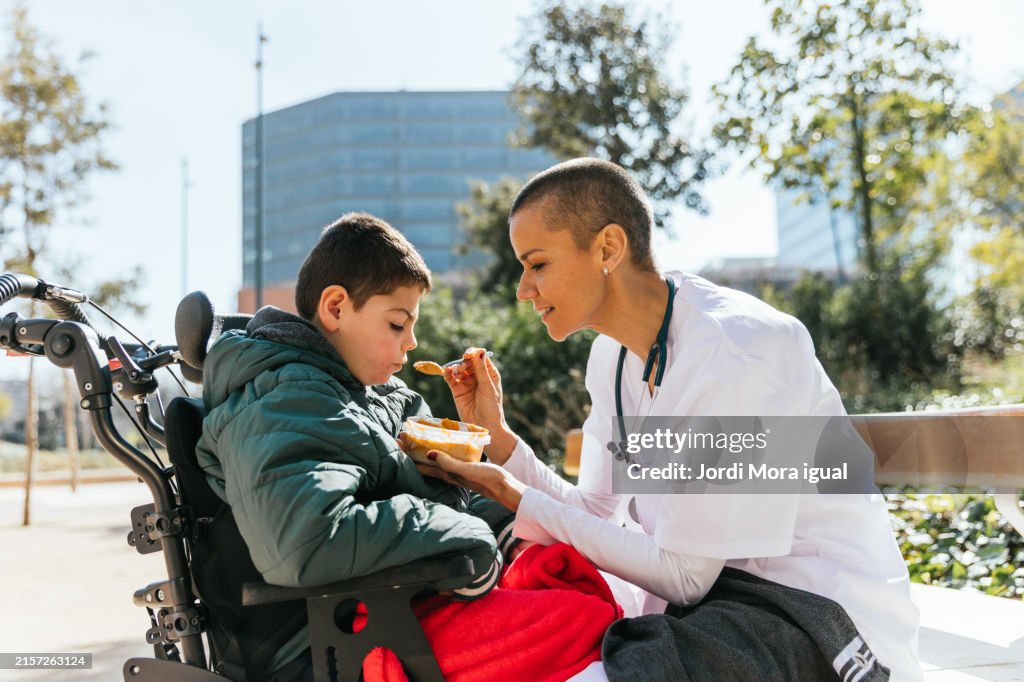 Young boy sits in a wheelchair in a park while a caregiver feeds him with a spoon.