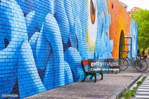 side view of a bench on the street sidewalk with a multi-color painted mirage on the wall with no people around - painted walls stock pictures, royalty-free photos & images