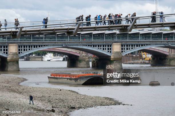 With Blackfriars Bridge in the distance tourists cross the Millennium Bridge over the River Thames as a lone figure walks along the foreshore at low...