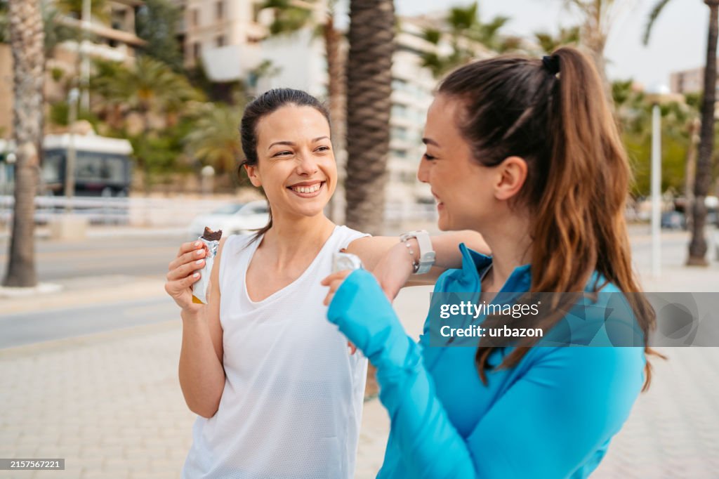 Two Female Friends Eating A Protein Bar After Training On The Quayside In Palma De Mallorca In Spain