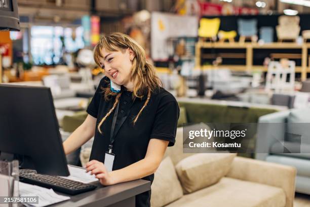 una vendedora sonriente trabaja en una computadora en una tienda departamental, contesta un teléfono fijo y habla con un cliente - teléfono con cable fotografías e imágenes de stock