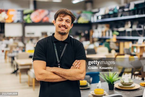 a handsome salesman in a furniture store looks directly into the camera while standing in the furniture store, surrounded by dining tables - loja de móveis imagens e fotografias de stock