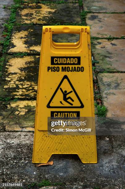 spanish- and english-language freestanding portable plastic warning sign stating 'cuidado piso mojado/caution wet floor' on a footpath at yaxhá archeological site, flores, petén, guatemala - opgepast-gladde-vloer stockfoto's en -beelden
