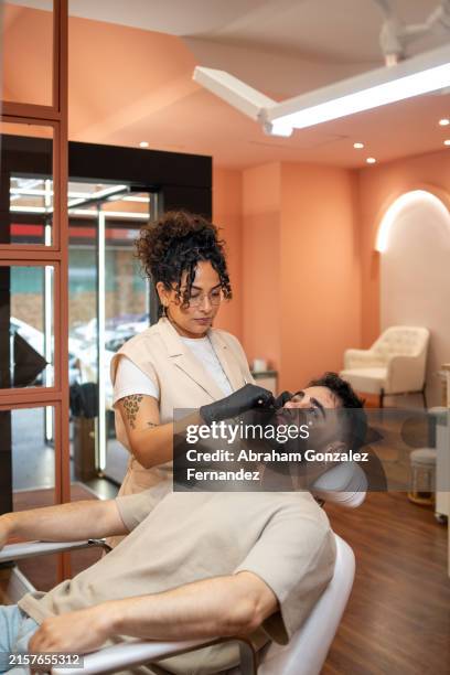 beautician wearing black gloves is using the threading technique to shape the eyebrows to a male customer in a beauty salon - threading eyebrows stock pictures, royalty-free photos & images