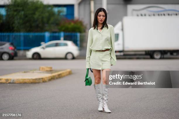 Cristine Sun wears a necklace, a pale pastel green oversize long shirt worn as a mini dress, a belt, a bold green leather Jacquemus bag, knee high...