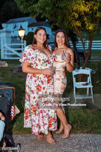 schöne puerto-ricanische erwachsene schwestern, die getränke in der hand halten und gemeinsam posieren, während sie nachts an der hochzeitsfeier ihres bruders im freien teilnehmen. - puertoricanischer abstammung stock-fotos und bilder