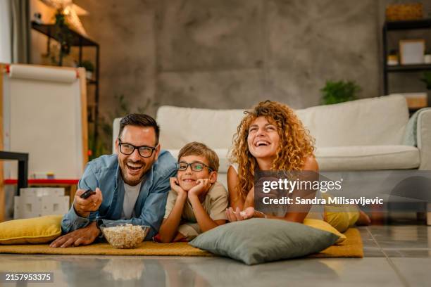 famiglia: padre, madre e figlio che si divertono a casa seduti sul pavimento a mangiare popcorn mentre guardano la tv - famiglia casa foto e immagini stock