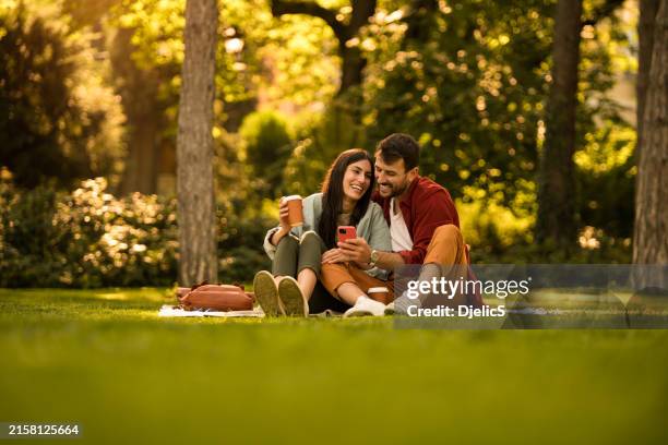 happy young couple using phone while relaxing together at a park on summer day. - couple picnic stock pictures, royalty-free photos & images