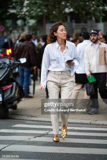 Guest wears silver necklace, light blue white lined button up long sleeve shirt, beige form fitted belted pants, shiny gold loafer leather shoes,...
