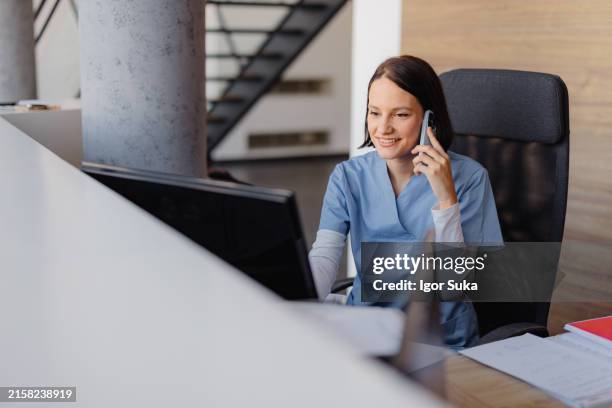 giovane receptionist donna che parla al telefono in clinica mentre è seduta e guarda sul monitor del pc - clinica medica foto e immagini stock