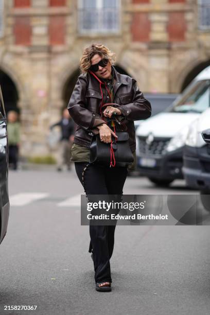 Guest wears black sunglasses, shiny dark brown leather jacket, dark green jacket, black Delvaux leather bag, a ring, black tight pants with...