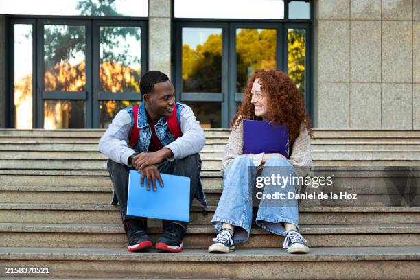 two happy multiracial university student friends enjoying break from classes laughing together. young friends sitting on stairs outside in college campus. - male college student sitting on campus steps stock pictures, royalty-free photos & images