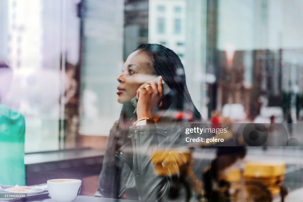 Porträt einer schwarzen Frau, die hinter der Fensterscheibe im Straßencafé mit dem Handy spricht