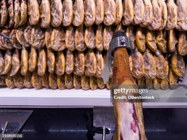a cured ham in the foreground, ready to cut, with a wall full of hams hanging in the background, showing hams in the background. concept of charcuterie, iberian, gourmet, delicatessen and cold cuts. - salty snack stock pictures, royalty-free photos & images