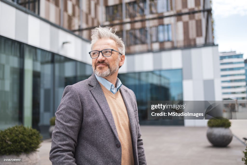 Reifer Geschäftsmann in einer städtischen Umgebung, mit Brille und Bart. In Blazer und Pullover gekleidet, strahlt er Selbstvertrauen und Professionalität aus. Urbane Szene mit modernen Gebäuden im Hintergrund.