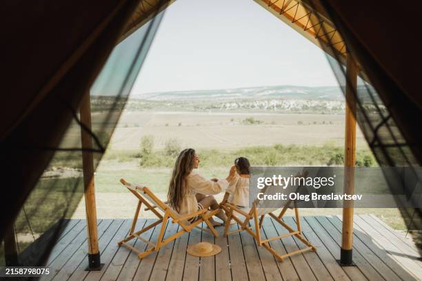 mom resting with daughter in chairs on wooden hotel terrace. view from behind. - glamping stock-fotos und bilder