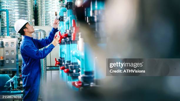 wartungsingenieur arbeiter mit schutzhelm, der ventile einstellt und manometer an industriellen rohrleitungen in einer wasseraufbereitungsanlage prüft. abfüllung von wasser - destilliertes wasser stock-fotos und bilder