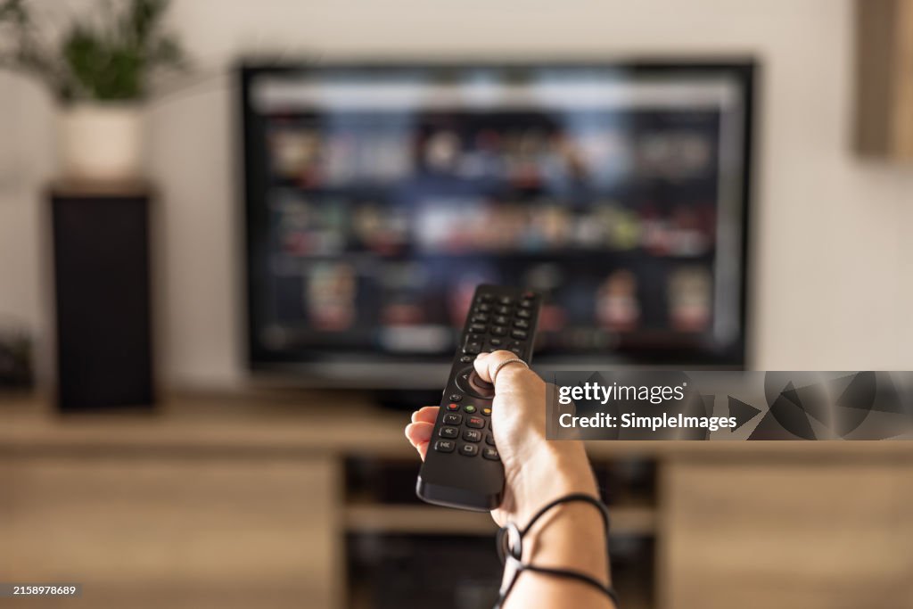 A woman is holding a remote control of a smart TV in his hand. In the background you can see the television screen with streaming entertainment apps for video on demand.